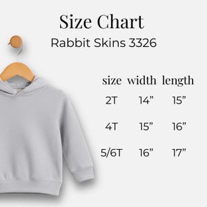 May include: A light gray hooded sweatshirt hanging on a wooden hanger. The image includes a size chart for Rabbit Skins 3326, with measurements in inches for sizes 2T, 4T, and 5/6T. The chart lists width and length.