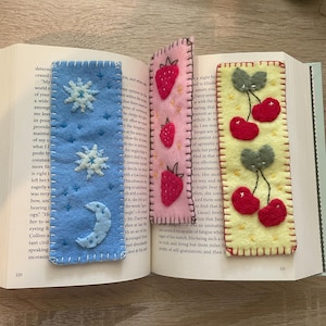 May include: Three rectangular felt bookmarks with hand-stitched edges resting inside an open book. The blue bookmark features white stars and a crescent moon, the pink one displays red strawberries, and the yellow one shows red cherries.