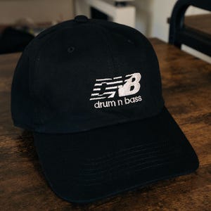 May include: Black baseball cap with a white New Balance logo and the text "drum n bass" embroidered on the front.