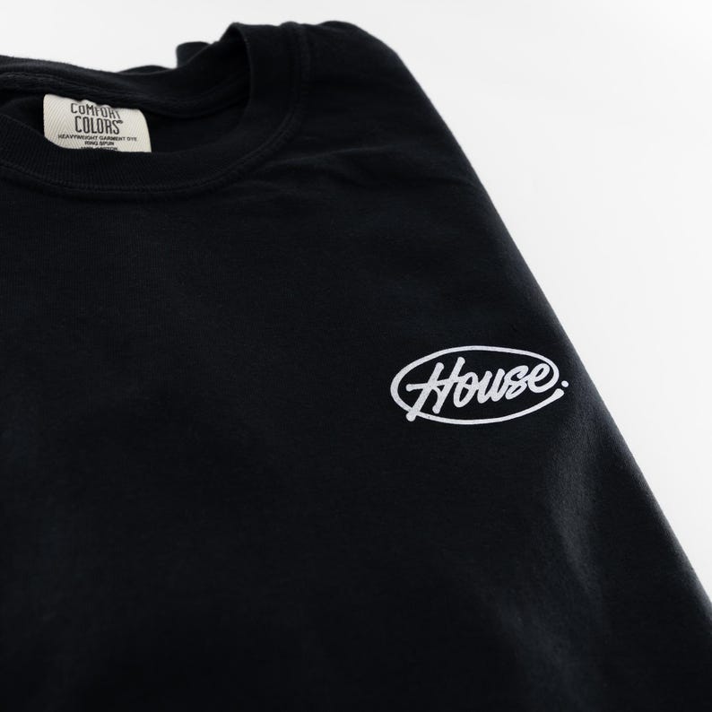 May include: A black crew neck t-shirt featuring a white oval graphic with the word House written in a script font. The interior neck label displays the text Comfort Colors, Heavyweight Garment Dye, Ring-Spun, and other small printed details.