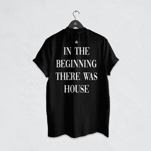 May include: A black short-sleeved t-shirt hanging on a black hanger. The back of the shirt features white text that reads IN THE BEGINNING THERE WAS HOUSE, with a small logo symbol positioned directly above the text on the upper back area.