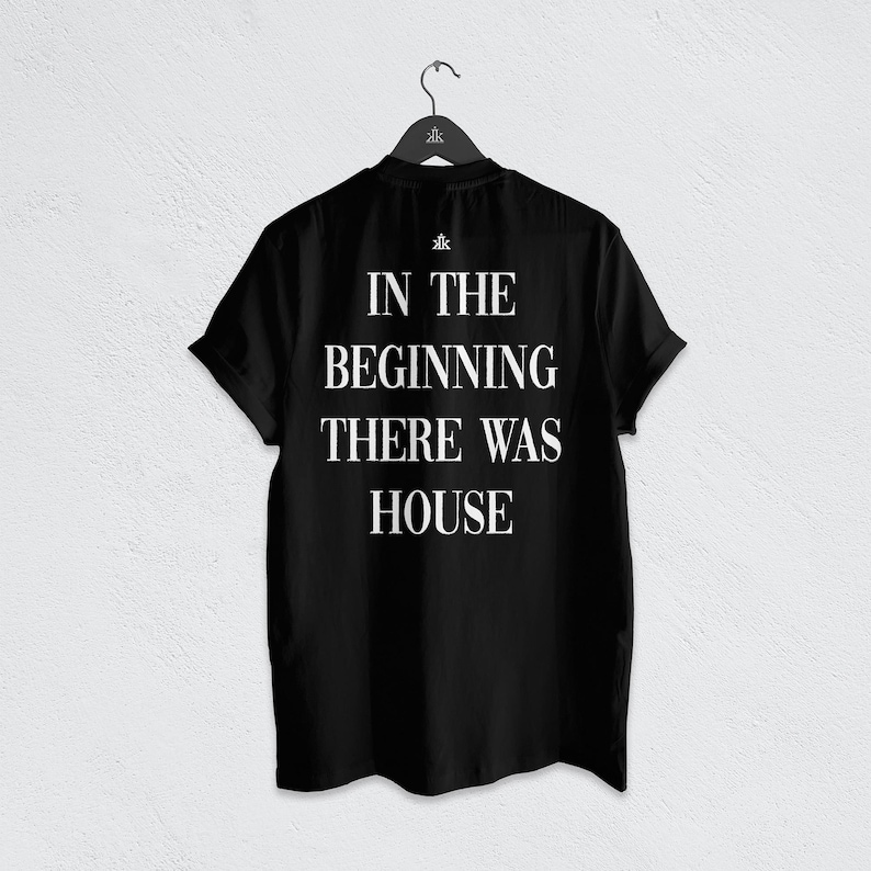 May include: A black short-sleeved t-shirt hanging on a black hanger. The back of the shirt features white text that reads IN THE BEGINNING THERE WAS HOUSE, with a small logo symbol positioned directly above the text on the upper back area.