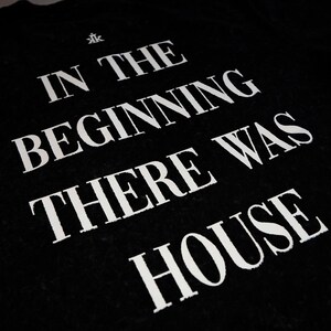 May include: A black t-shirt featuring white text that reads IN THE BEGINNING THERE WAS HOUSE, with a small white logo above the text. The lettering is printed in a bold, serif font, creating a high-contrast design against the dark fabric background.