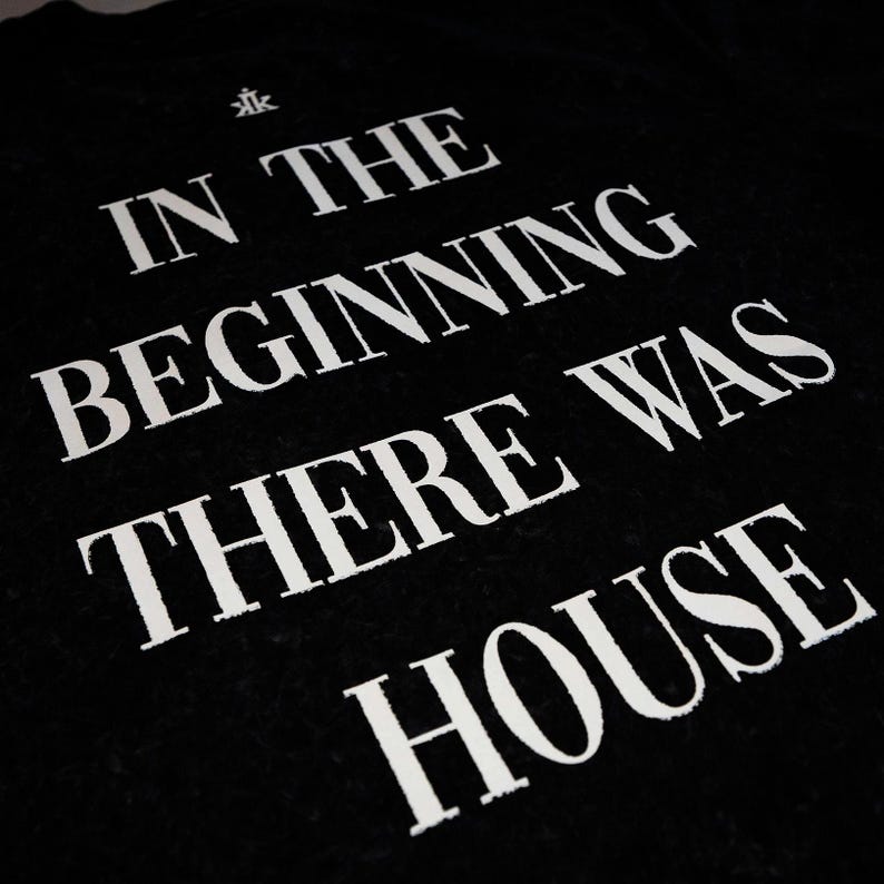 May include: A black t-shirt featuring white text that reads IN THE BEGINNING THERE WAS HOUSE, with a small white logo above the text. The lettering is printed in a bold, serif font, creating a high-contrast design against the dark fabric background.