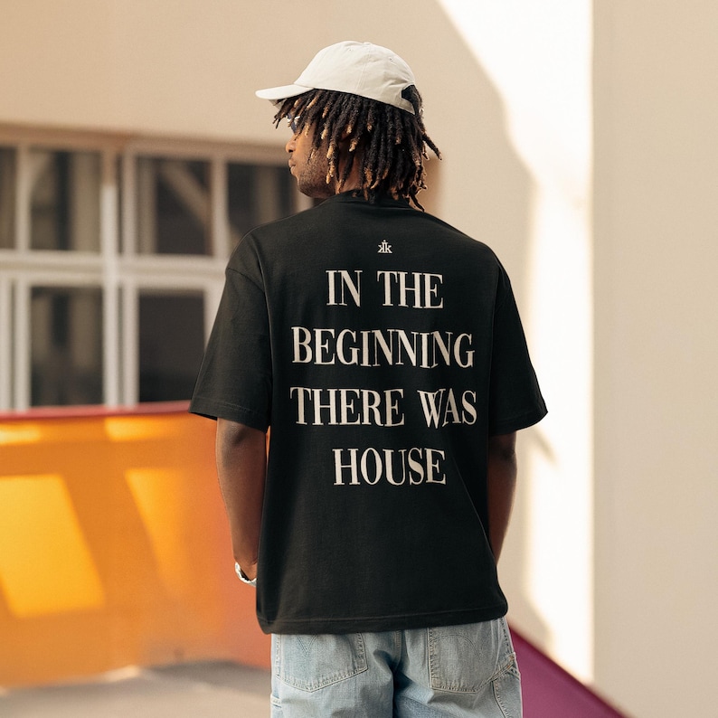 May include: A black short-sleeve t-shirt featuring the white text "IN THE BEGINNING THERE WAS HOUSE" printed on the back, accompanied by a small white logo above the text. The shirt is paired with a light-colored baseball cap and blue jeans.