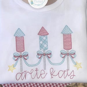 Könnte beinhalten: Weißer Baby-Body mit gestickten Feuerwerkskörpern in Rosa und Blau, verbunden durch eine hellblaue Naht. Der Text "are te kats" ist darunter in roter Schrift gestickt. Ein Etikett mit dem Text "Once Upon a Thread" ist angebracht.