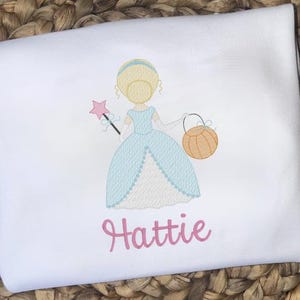 May include: White garment with an embroidered princess design. The princess is in a light blue dress, holding a pink star wand and an orange pumpkin basket. The name "Hattie" is written in pink script below the princess.