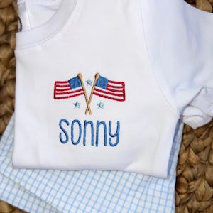 Könnte beinhalten: Ein weißes kurzärmeliges T-Shirt mit einer Stickerei aus zwei gekreuzten amerikanischen Flaggen, hellblauen Sternen und dem in blauem Garn gestickten Namen Sonny. Das T-Shirt liegt auf einem hellblau-weiß karierten Vichy-Stoff.