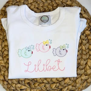 May include: A white short-sleeved t-shirt featuring an embroidered design of three round fish in light blue, pink, and lavender, each with a small bow and bubbles. The name Lilibet is embroidered in pink cursive script below the fish.