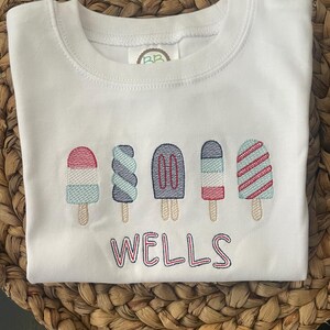 Könnte beinhalten: Weißes Sweatshirt mit Rundhalsausschnitt und gesticktem Eis am Stiel-Design in Rot, Blau und Türkis. Unterhalb der Eis am Stiel ist der Schriftzug "WELLS" gestickt. Präsentiert auf einer braunen, gewebten Oberfläche.