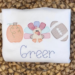 Può includere: T-shirt bianca con ricami a tema autunnale. Presenta una zucca, un tacchino e un pallone da football in arancione, rosa, blu e marrone. Il nome "Greer" è ricamato in blu sotto le immagini.