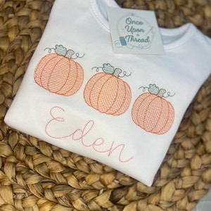 May include: White long-sleeved shirt featuring three embroidered orange pumpkins with green stems. The name "Eden" is embroidered in pink script. A tag with "Once Upon a Thread" is attached.