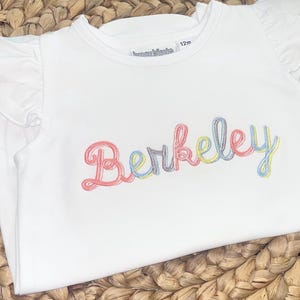 Puede incluir: Camiseta blanca de bebé con mangas con volantes y el nombre "Berkeley" bordado en hilo colorido. La camiseta es talla 12m y está hecha de un material suave. La camiseta está sobre una cesta tejida.