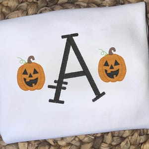 May include: White shirt with a large black letter "A" and two orange pumpkins with carved faces. The pumpkins have green vines and are a Halloween-themed design. The shirt is a festive item.
