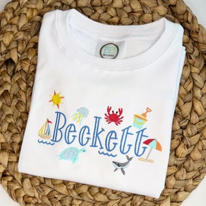 Op de afbeelding: Wit T-shirt met geborduurde strandthema's, waaronder een zeilboot, zon, krab en parasol. De naam "Beckett" is in blauwe letters geborduurd. Een klein logo is zichtbaar bij de halslijn.