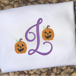 May include: White fabric with an embroidered Halloween design. It features a large purple letter "L" and two orange jack-o'-lanterns with green stems and smiling faces. The design is centered on the fabric, suitable for seasonal decor.
