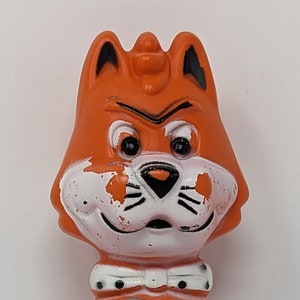 May include: Orange plastic figurine of a cat with a white face and black eyes. The cat is holding two gray mice in its arms. The cat is wearing a white collar with black dots and a white bow tie. The text "MR. JINKS PIXIE AND DIXIE" is printed on the bottom of the figurine.