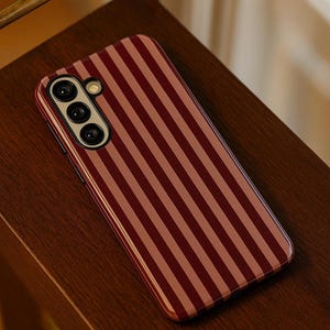 Op de afbeelding: Een gestreepte telefoonhoes in bordeauxrood en lichtrood. De hoes ligt op een houten oppervlak. De telefoonhoes heeft een camera met drie lenzen. De hoes is ontworpen om de telefoon te beschermen tegen krassen en beschadigingen.