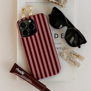 Könnte beinhalten: Eine Sammlung von Accessoires, darunter eine gestreifte Handyhülle, Sonnenbrille, Haarspangen, Ohrringe und eine Tube Rhode-Produkt. Die Handyhülle hat ein rosa-burgunderfarbenes vertikales Streifenmuster. Die Sonnenbrille hat einen Schildpattrahmen.