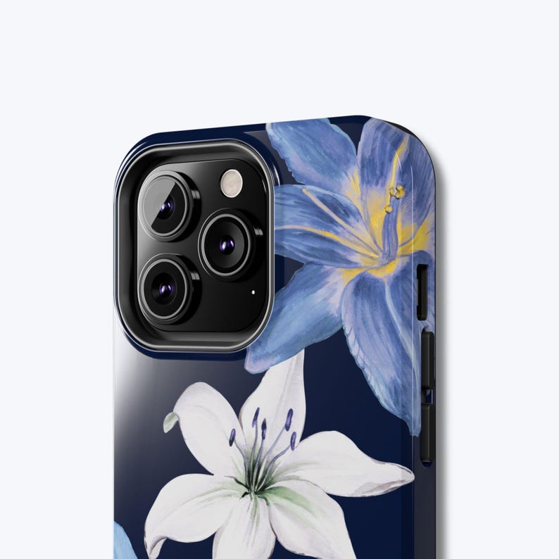 May include: A navy blue phone case with a floral pattern of white and blue lilies. The case has a glossy finish and a camera cutout with three lenses. The lilies have yellow centres and are set against a dark background.