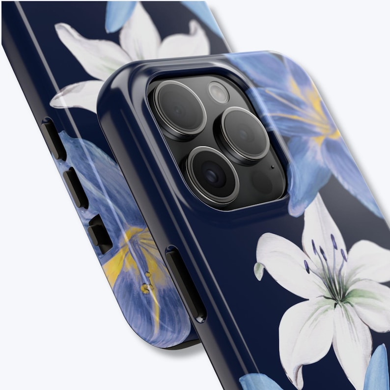 May include: A navy blue phone case with a floral pattern of white and blue lilies. The case has a glossy finish and a raised edge around the camera lenses. The design includes yellow accents within the flower petals.