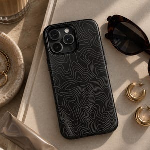 Può includere: Una custodia per smartphone nera con un motivo a linee topografiche bianche. La custodia ha una forma rettangolare con angoli arrotondati e un'apertura per l'obiettivo della fotocamera sul lato superiore sinistro.
