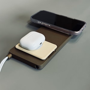 Peut inclure: Une station de charge marron avec un chargeur sans fil blanc pour téléphone et un chargeur sans fil blanc pour AirPods. Un étui de téléphone transparent est sur le chargeur de téléphone.