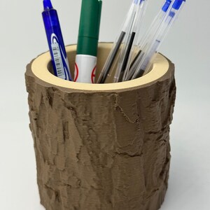 Peut inclure: Un porte-stylo marron avec un extérieur texturé en forme d'écorce. L'intérieur est de couleur beige clair. Le porte-stylo contient un stylo bleu, un marqueur vert et plusieurs stylos transparents à encre noire.