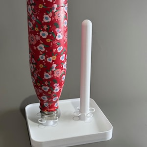 Puede incluir: Una botella de agua roja con estampado floral, con un soporte blanco y un accesorio cilíndrico blanco. La botella está decorada con un patrón repetido de flores rosas, azules y blancas. El soporte es cuadrado y tiene un borde elevado.
