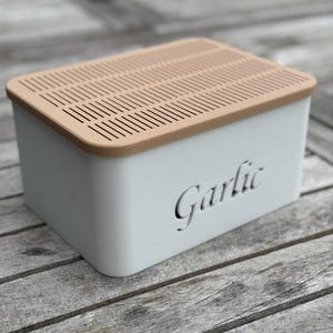 May include: White ceramic garlic keeper with a light brown lid featuring a grid pattern. The lid has a cutout that says "Garlic".