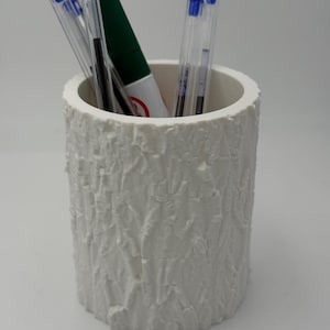 Peut inclure: Un pot à crayons blanc, cylindrique, avec une surface texturée ressemblant à de l'écorce. Il contient plusieurs stylos transparents à capuchons bleus et un marqueur vert. Un organisateur de bureau.