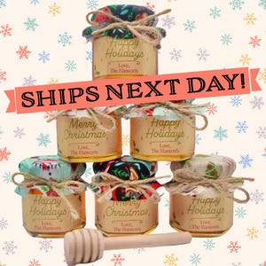 May include: Six small glass jars with festive Christmas and holiday themed labels. The jars are filled with honey and are tied with twine. A wooden honey dipper is in the foreground. The jars are arranged in a pyramid shape. The text on the labels reads "Happy Holidays" and "Merry Christmas".