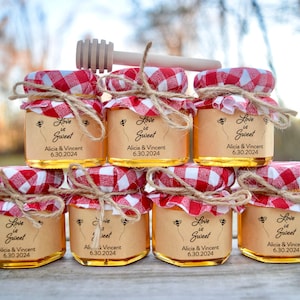 Puede incluir: Ocho pequeños frascos de vidrio llenos de miel, decorados con tela a cuadros rojos y blancos, cuerda y un pequeño amuleto de abeja. Cada frasco tiene una etiqueta que dice "Love is Sweet" con los nombres Alicia & Vincent y la fecha 6.30.2024.