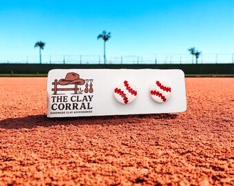 May include: Baseball stud earrings with red stitching on a white card. The card reads "THE CLAY CORRAL HANDMADE CLAY ACCESSORIES". The earrings are displayed on a baseball field with a blue sky in the background.