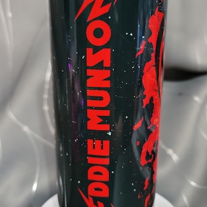 Pode incluir: Um copo de aço inoxidável preto e vermelho com um design de respingo preto e branco. O copo tem um raio vermelho e o texto "EDDIE MUNSON" em vermelho.