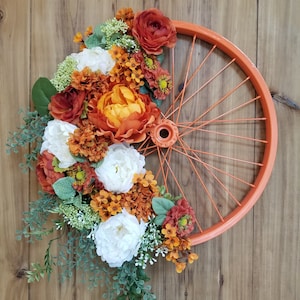 May include: A bright orange painted bicycle wheel with a floral wreath made of white and orange flowers. The wreath is attached to the wheel with wire.