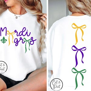 Könnte beinhalten: Weißes Sweatshirt mit dem Text "Mardi Gras" in Lila und Gelb, mit einer grünen Fleur-de-Lis. Auf der Rückseite befinden sich drei Schleifen in Gelb, Lila und Grün. Das Sweatshirt ist von "Cut & Print Shop".