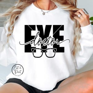 Puede incluir: Sudadera blanca con la frase "EYE doctor" en un diseño gráfico negro, con gafas debajo del texto. La sudadera tiene mangas largas y cuello redondo. El logotipo "Cat & Print SVG Shop" está en la parte inferior.