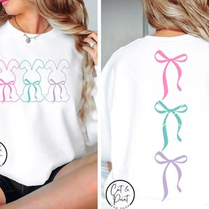 May include: White sweatshirt featuring three bunny silhouettes with pink, teal, and purple bows on the front. The back of the sweatshirt displays three matching bows. The text "Cut & Print Shop" is visible on the bottom left.