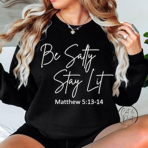 以下が含まれることがあります： 白い文字で「Be Salty Stay Lit」と「Matthew 5:13-14」と書かれた黒いスウェットシャツ。スウェットシャツはクルーネックと長袖です。画像には「Cat & Print SVG Shop」のロゴも含まれています。