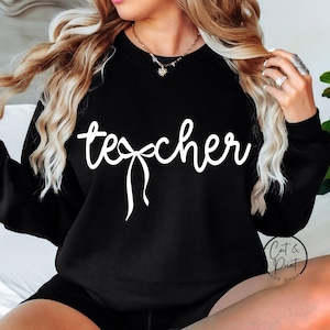 May include: Black sweatshirt with the word "teacher" in white cursive script, featuring a bow design. The sweatshirt is a long-sleeved crew neck. The image also shows a person with blonde hair.