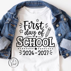 May include: A white t-shirt featuring black text that reads First day of SCHOOL 2026-2027, decorated with a pencil icon, stars, and small hearts. The shirt is layered underneath a blue denim jacket with metal buttons and folded cuffs.