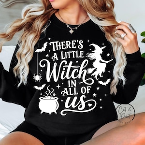 May include: A black long-sleeve sweatshirt featuring white graphic text that reads There's A Little Witch In All Of Us. The design includes illustrations of a witch on a broomstick, a bubbling cauldron, a spider, bats, and small stars.