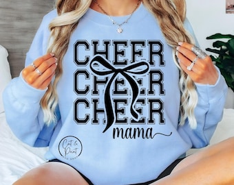 Może przedstawiać: Jasno niebieski sweter z czarnym napisem "Cheer Cheer Cheer mama" i czarnym graficznym motywem kokardki.