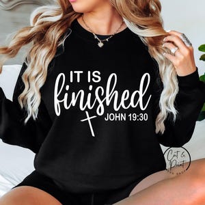 May include: Black sweatshirt with the text "IT IS finished" in white script, with a cross and the text "JOHN 19:30" below. The sweatshirt has long sleeves and a crew neck. The image also includes the logo "Cat & Print Shop".