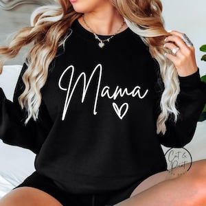 Könnte beinhalten: Schwarzes Sweatshirt mit dem Wort "Mama" in weißer Schrift und einem kleinen Herz. Das Sweatshirt hat einen Rundhalsausschnitt. Das Logo von Cat & Print SVG Shop ist unten rechts sichtbar.