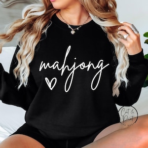 May include: Black sweatshirt with the word "mahjong" in white script, accompanied by a small heart. The sweatshirt is part of the "Cat & Print SVG Shop" collection.