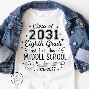 May include: A white t-shirt featuring black text that reads Class of 2031 Eighth Grade last first day of MIDDLE SCHOOL 2026-2027, accompanied by a graphic of a smiley face and a pencil, layered under a blue denim jacket with metal buttons.