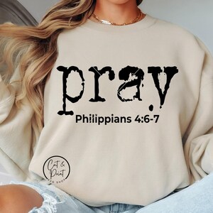 Puede incluir: Sudadera beige con la palabra "pray" en fuente negra desgastada. Debajo está el texto "Philippians 4:6-7". La sudadera tiene cuello redondo y mangas largas.