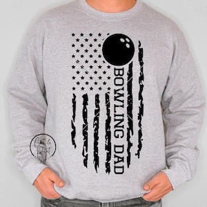 May include: A light gray sweatshirt featuring a distressed American flag design with a bowling ball and the words "Bowling Dad" in black. The sweatshirt has long sleeves and a crew neck.